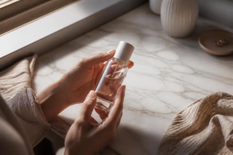 Hands holding a minimalist glass skincare bottle on a marble counter in soft natural light