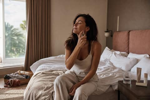 Woman resting a chilled mist bottle against her cheek in a sunlit hotel room beside a half-packed bag