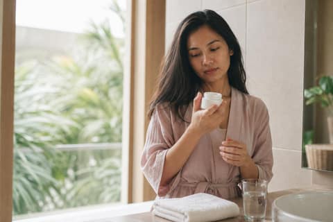 A woman examining a small skincare jar at a sunlit bathroom counter in a modern Malaysian apartment