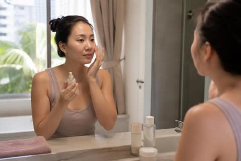 A woman in a bright Singapore bathroom holding a small serum bottle and checking her skin in the mirror.