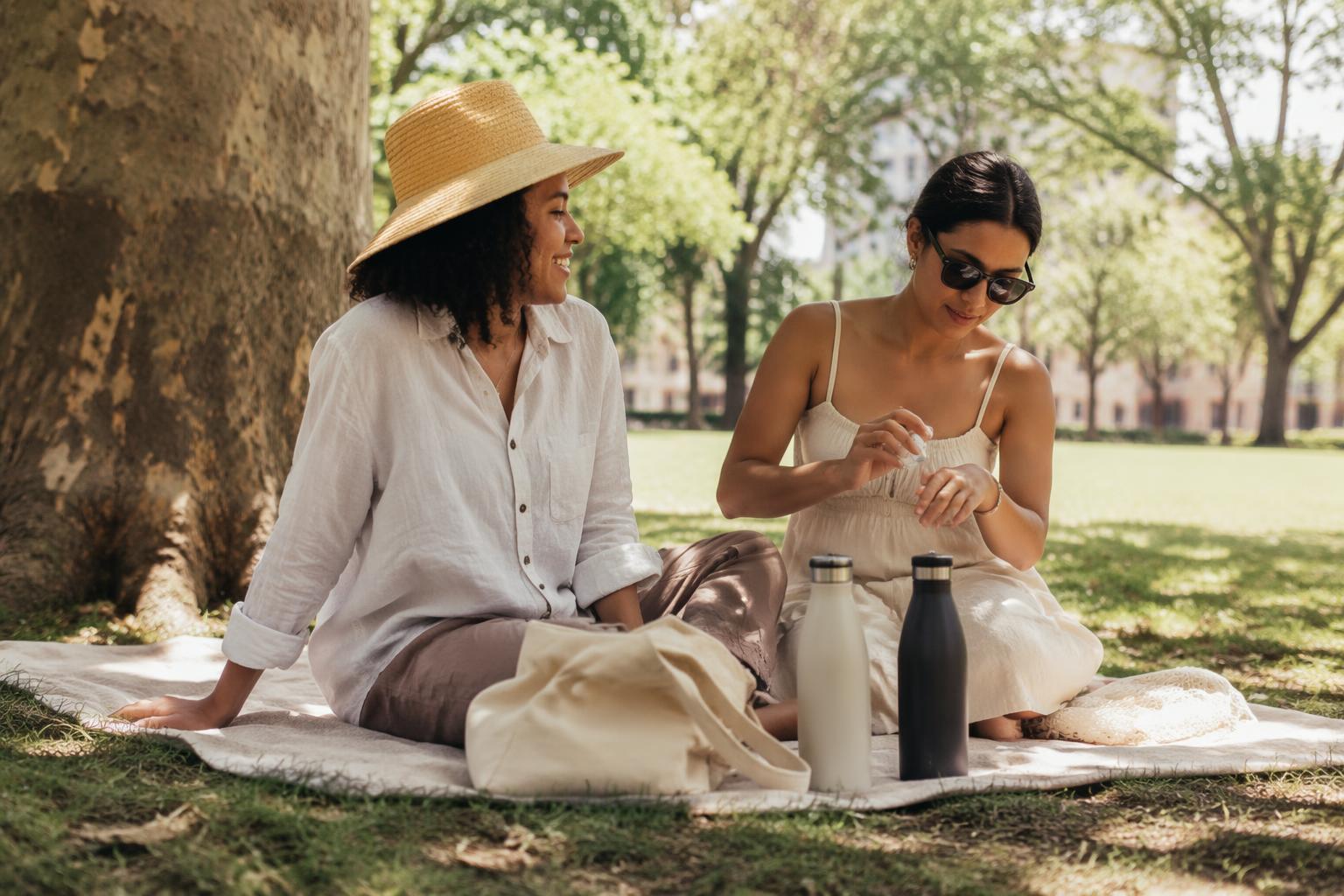 Two friends relaxing in tree shade in a sunny city park, one wearing a wide-brimmed hat, the other applying lotion to her hand