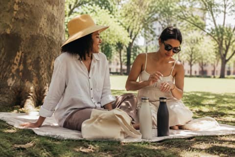 Two friends relaxing in tree shade in a sunny city park, one wearing a wide-brimmed hat, the other applying lotion to her hand