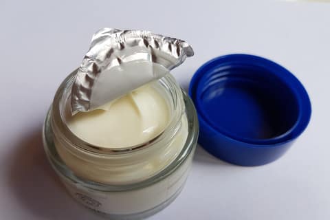 An open jar of face cream with the foil seal peeled back beside its blue lid